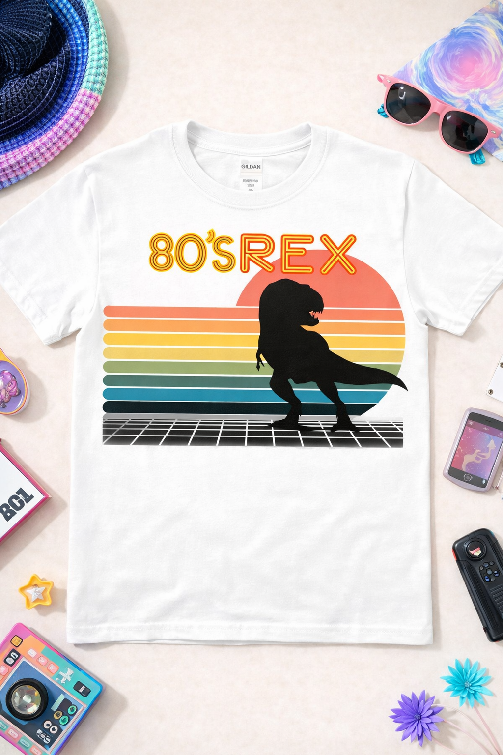 White graphic t-shirt featuring “80’s REX” retro dinosaur design with colorful sunset stripes and T-Rex silhouette, styled in a flatlay with 80s inspired accessories and props