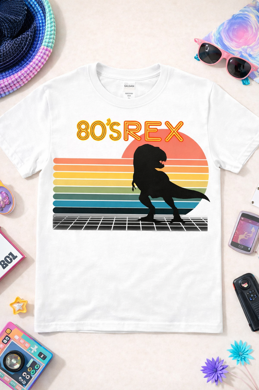 White graphic t-shirt featuring “80’s REX” retro dinosaur design with colorful sunset stripes and T-Rex silhouette, styled in a flatlay with 80s inspired accessories and props