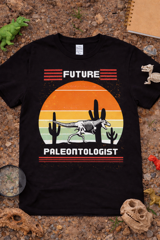 Black graphic t-shirt featuring white T-Rex skeleton with retro sunset desert and cactus design reading “Future Paleontologist,” styled in flat lay with dinosaur props