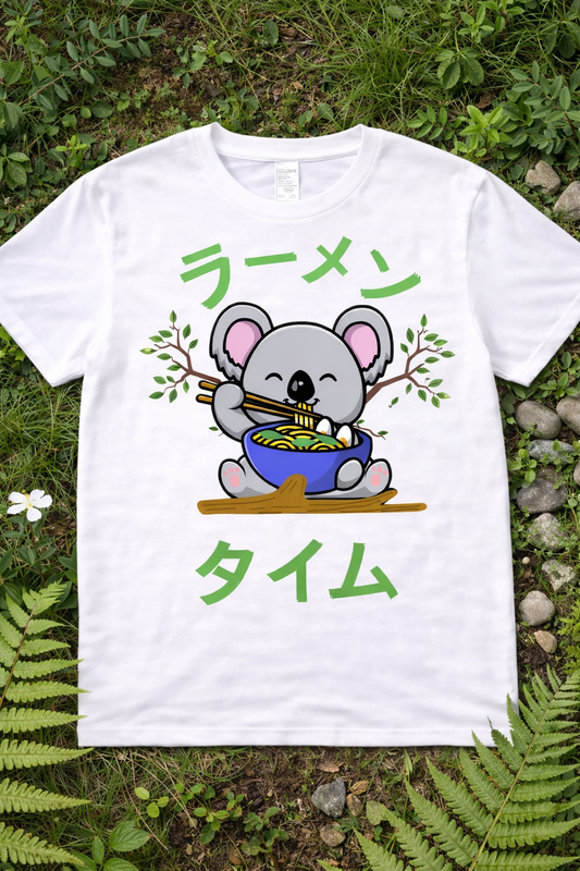 Kawaii koala ramen graphic tee on white shirt featuring cute cartoon koala eating noodles with chopsticks, Japanese ramen text, anime foodie design flatlay on grass with nature background