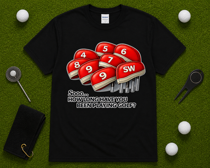 Black funny golf shirt flatlay featuring How Long Have You Been Playing Golf graphic with golf headcovers and golf accessories on putting green backdrop. Golf lover gift shirt for tournament players, golf coaches, golf retirees, golf fans, and casual golfers across the United States.
