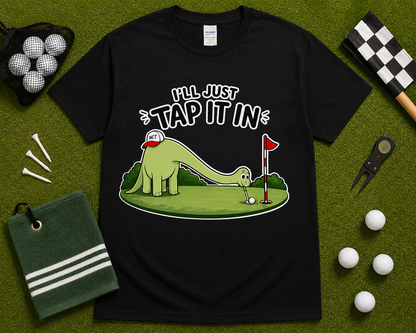 Black funny golf dinosaur shirt flatlay featuring Ill Just Tap It In brontosaurus golfer design on golf themed grass background golf gift for men women and youth USA