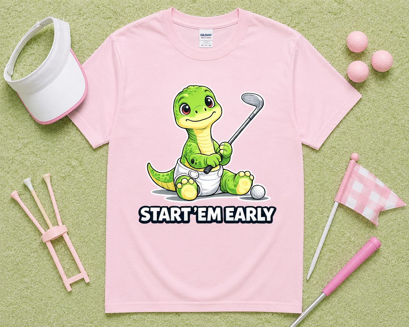 Light Pink Gildan 5000 Start Em Early dinosaur golf shirt flatlay with golf accessories on light green grass, funny golf graphic tee gift for girls, moms, junior golfers and golf families in the USA.