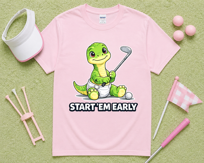 Light Pink Gildan 5000 Start Em Early dinosaur golf shirt flatlay with golf accessories on light green grass, funny golf graphic tee gift for girls, moms, junior golfers and golf families in the USA.