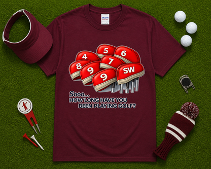 Maroon golf shirt flatlay featuring funny How Long Have You Been Playing Golf graphic with red golf headcovers and golf accessories. Fun golf apparel for golfers, golf league players, tournament participants, golf dads, and golf gift buyers throughout California, Texas, Florida, and the United States.