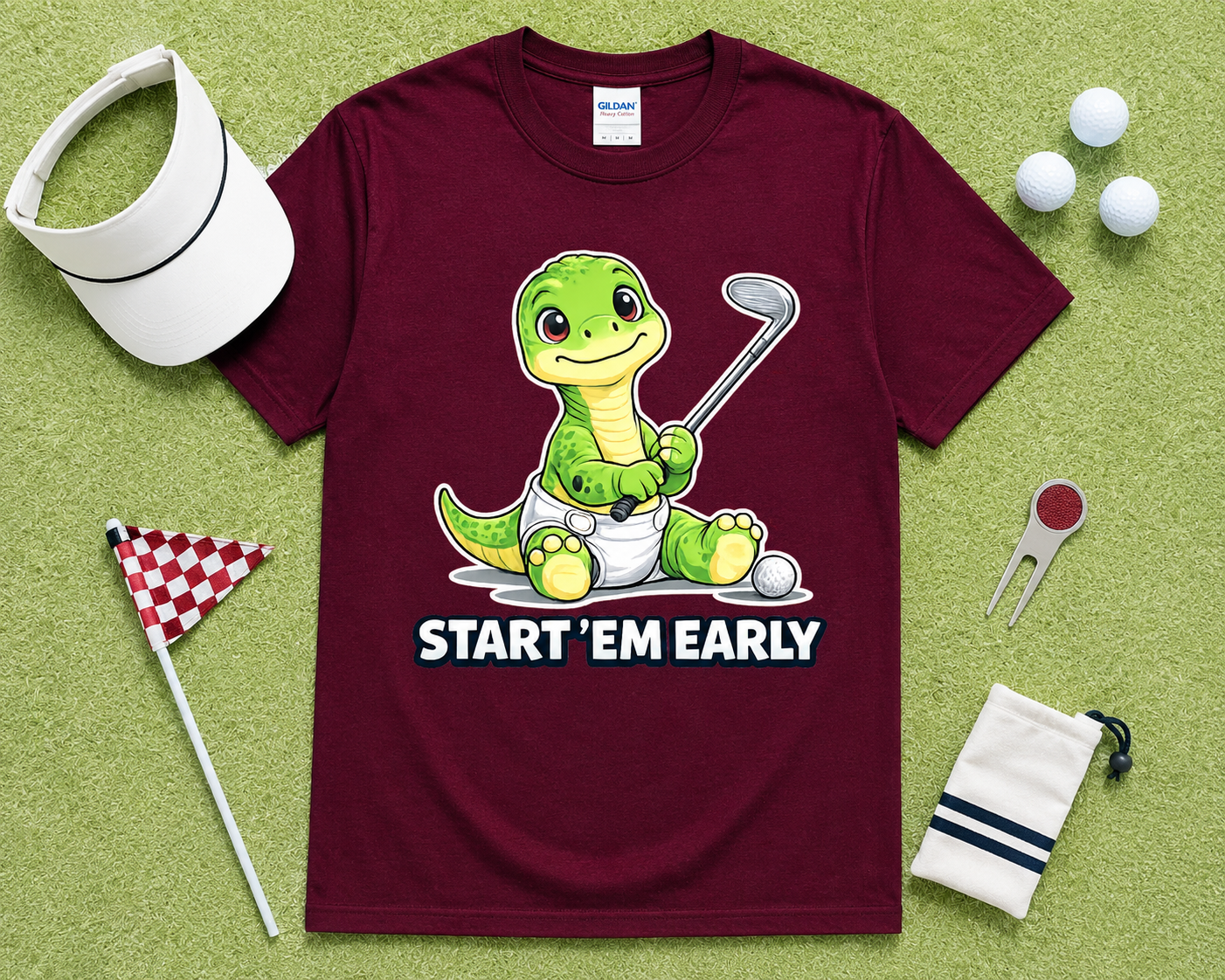 Maroon Gildan 5000 funny Start Em Early dinosaur golf shirt flatlay with golf accessories on light green grass, cute golf graphic tee gift for golfers, junior golfers and golf families in the USA.