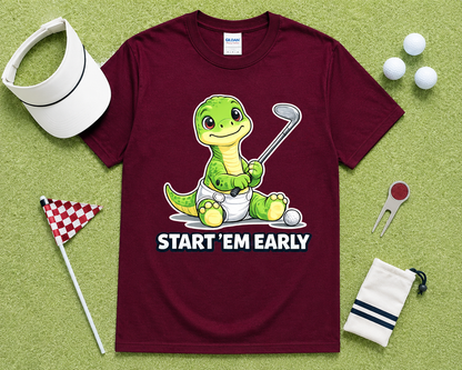Maroon Gildan 5000 funny Start Em Early dinosaur golf shirt flatlay with golf accessories on light green grass, cute golf graphic tee gift for golfers, junior golfers and golf families in the USA.