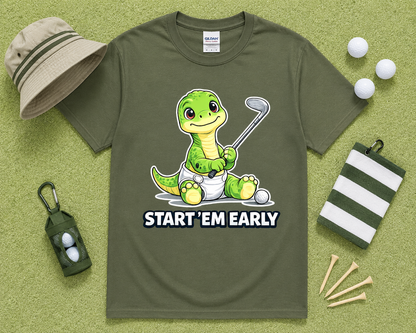 Military Green Gildan 5000 funny dinosaur golf shirt flatlay with golf accessories on light green grass, Start Em Early golf graphic tee for golfers, dads and golf families throughout the USA.