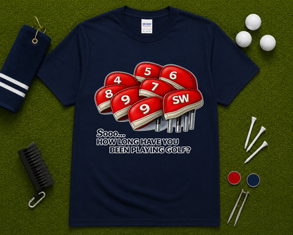 Navy golf graphic tee flatlay showcasing funny How Long Have You Been Playing Golf design with golf club headcovers and golf themed accessories. Popular golf lifestyle shirt for men and women, golf trips, golf tournaments, golf outings, and golf gift occasions nationwide.