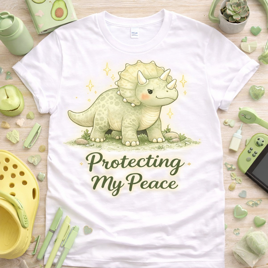 Cute triceratops shirt with Protecting My Peace text on a white t shirt flatlay with soft green aesthetic props cozy calm lifestyle outfit