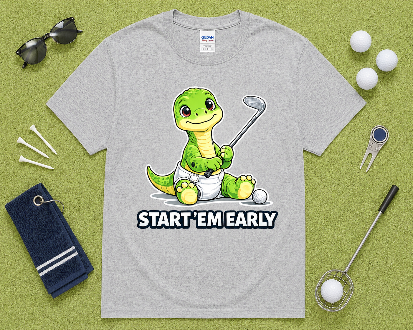 Sport Gray Gildan 5000 cute dinosaur golf shirt flatlay with golf accessories on light green grass, funny Start Em Early golf graphic tee for golfers, dads, moms and families in the USA.