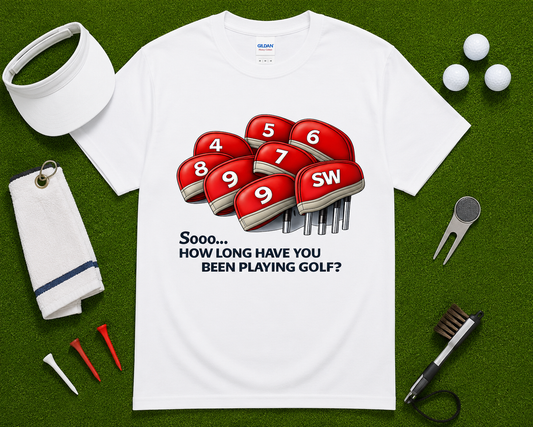 White golf shirt flatlay with funny How Long Have You Been Playing Golf graphic featuring numbered golf club headcovers and golf accessories on putting green background. Great golf gift for men, women, golf dads, golf coaches, golf retirees, and weekend golfers across the United States.