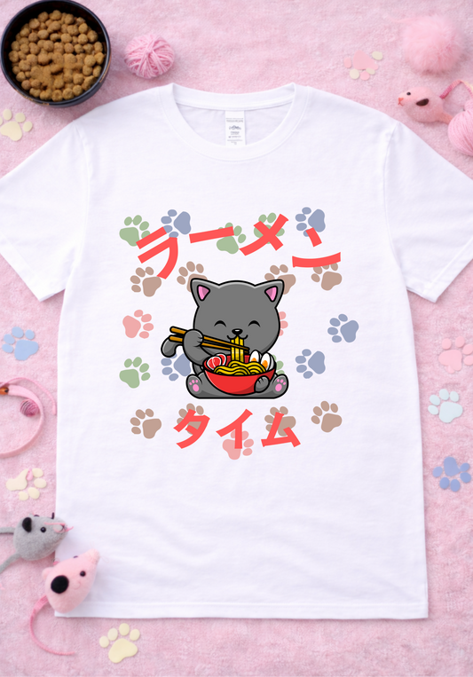 Kawaii gray cat ramen graphic tee on white shirt featuring cute cartoon cat eating noodles with chopsticks, colorful paw prints, Japanese ramen text, anime foodie design flatlay with cat toys and yarn