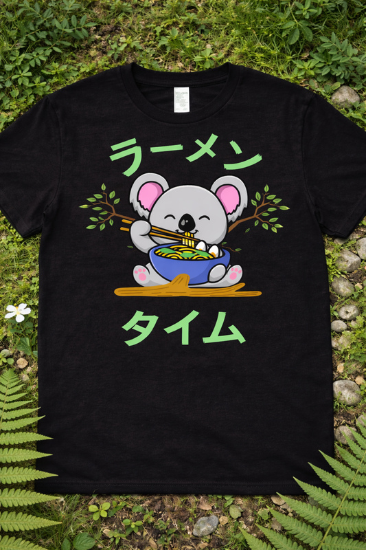 Kawaii koala ramen graphic tee on black shirt featuring cute cartoon koala eating noodles with chopsticks, Japanese ramen text, anime foodie design flatlay on grass with nature background