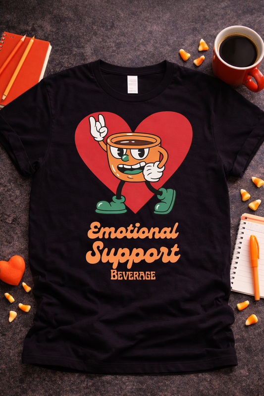 Black graphic t-shirt featuring retro coffee cup character with red heart and text “Emotional Support Beverage,” styled in flat lay with coffee mug, notebook, and warm-toned accessories
