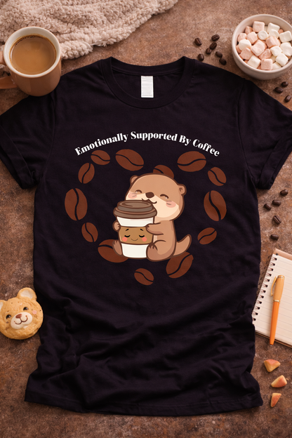 Black graphic t-shirt featuring cute otter hugging coffee cup with coffee beans and text “Emotionally Supported by Coffee,” styled in cozy flat lay with latte, marshmallows, and warm neutral props