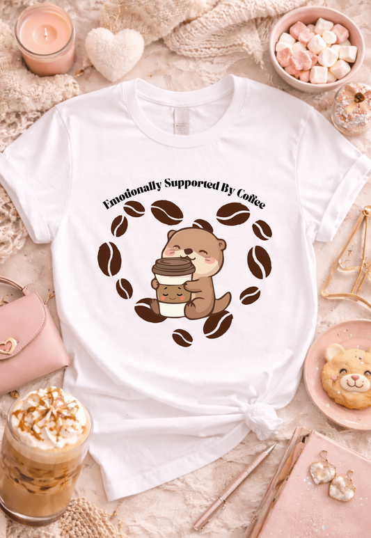 White graphic t-shirt featuring “Emotionally Supported By Coffee” otter design in a cozy flatlay with iced coffee, marshmallows, candle, and bear cookie, kawaii coffee aesthetic outfit