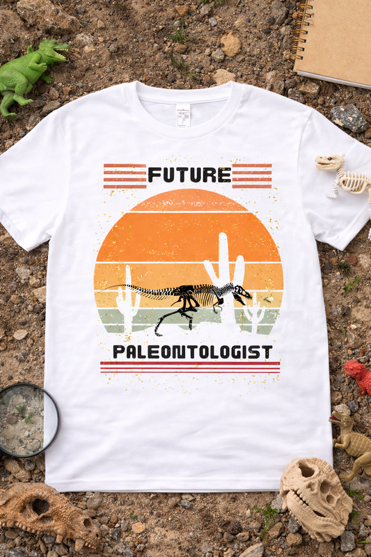White graphic t-shirt featuring “Future Paleontologist” text with dinosaur skeleton and vintage desert sunset design, styled in a flatlay with fossils, rocks, and dinosaur toys