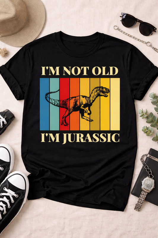 Black graphic t-shirt featuring “I’m Not Old I’m Jurassic” retro dinosaur design with colorful vintage stripes, styled in a flatlay with sneakers, sunglasses, and lifestyle props