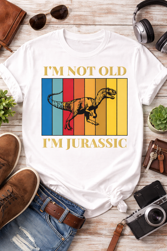 White graphic t-shirt featuring “I’m Not Old I’m Jurassic” retro dinosaur design with multicolor vintage stripes, styled in a flatlay with jeans, camera, and casual lifestyle props
