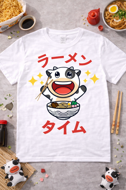 White graphic t-shirt featuring kawaii cow eating ramen with sparkles and Japanese text, styled in a clean flatlay with ramen bowls and themed accessories