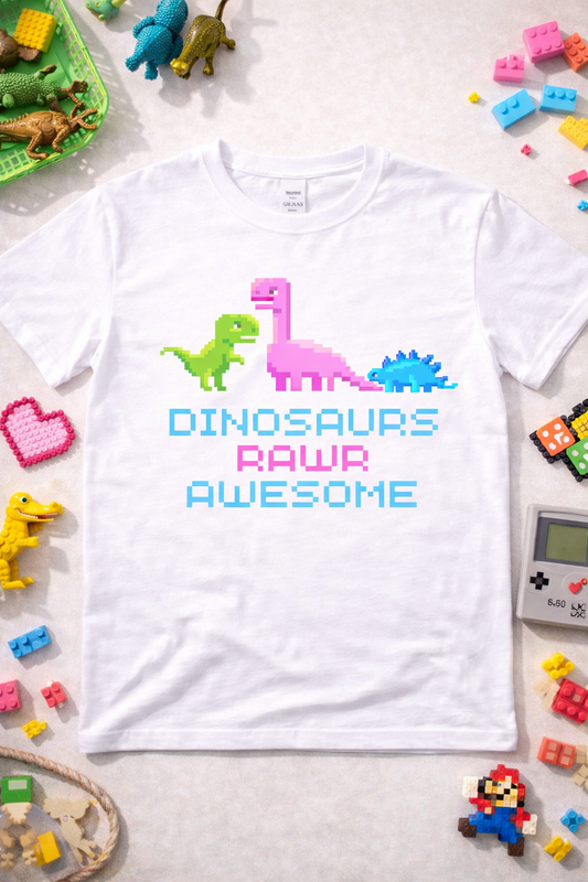 White graphic t-shirt featuring pixel dinosaurs and “Dinosaurs Rawr Awesome” text in 8-bit retro style, styled in a playful flatlay with colorful toys and gaming accessories
