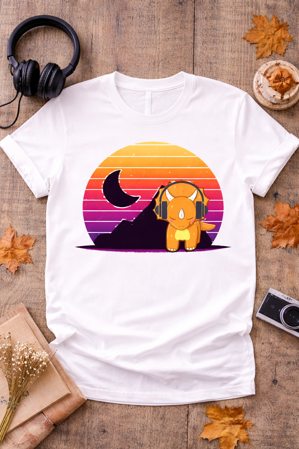 White graphic t-shirt featuring retro triceratops with headphones and vintage sunset design styled in cozy flat lay with headphones, camera, and autumn decor