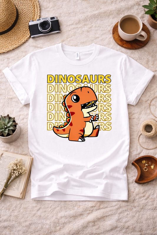 White graphic t-shirt featuring a cute retro T-Rex dinosaur design with repeating “Dinosaurs” text, styled in a neutral flatlay with coffee, camera, and cozy lifestyle props