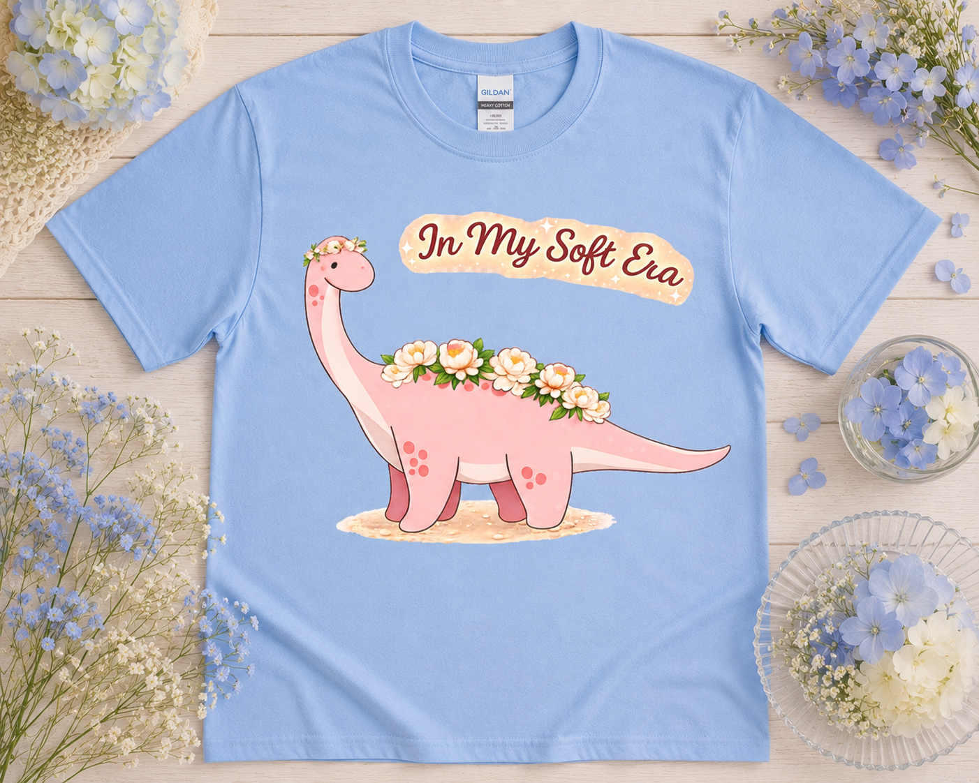 Cute pastel dinosaur graphic tee on Carolina blue Gildan shirt with dreamy floral styled flatlay