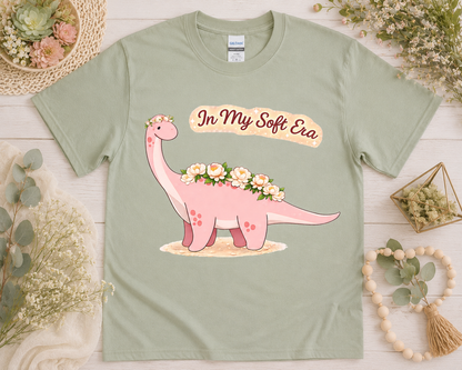 Floral brontosaurus kawaii graphic tee on sage green Gildan shirt with soft botanical flatlay props